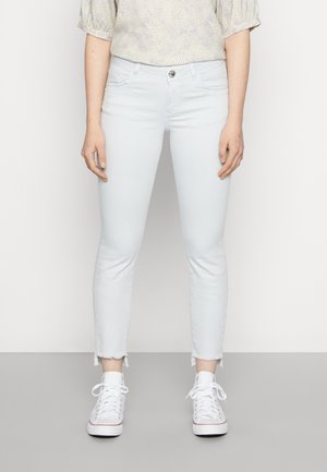 Jeans ajustados de color azul claro con dobladillo deshilachado. Hechos de denim, cuentan con cinco bolsillos y cierre con botón en la parte delantera. Llevados con zapatillas blancas.