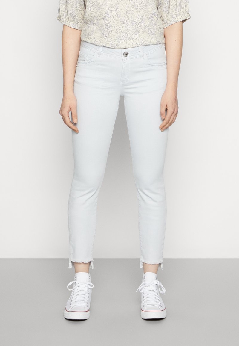 Světle modré, úzké džíny s roztrženým lemem. Vyrobené z denimu, obsahují pět kapes a zapínání na přední knoflík. Nohavice jsou obuty s bílými teniskami.