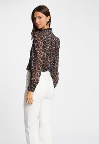 Chemisier en dentelle à manches longues en imprimé léopard marron. Caractéristiques : un col et des poignets à boutons. Assorti à un pantalon blanc taille haute.