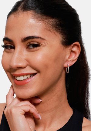Silberne Creolen mit kleinen, funkelnden Edelsteinen. Glatte, polierte Oberfläche, mit einem einfachen, kreisförmigen Design. Benutzerin mit leichtem Make-up.
