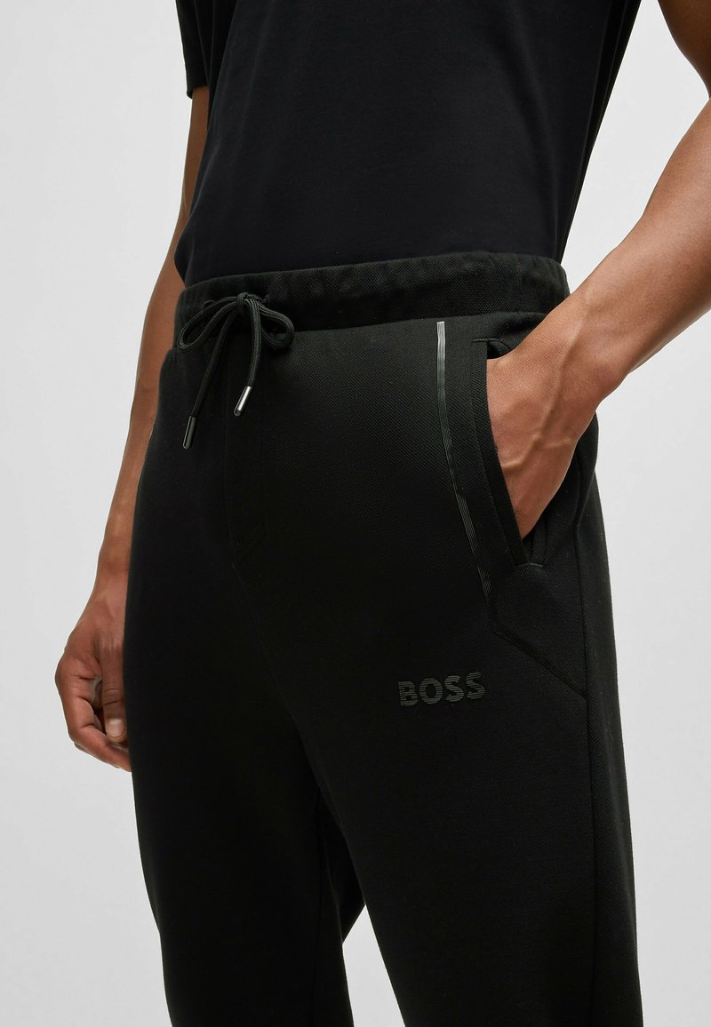 Pantalones jogger negros con tejido texturizado, cinturilla con cordón, bolsillos laterales y un sutil logo "BOSS" en relieve en la parte delantera.