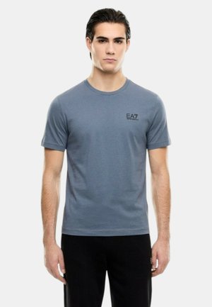 Giovane uomo con capelli scuri indossa una maglietta attillata blu con il marchio EA7 e pantaloni neri, in piedi davanti a uno sfondo chiaro uniforme.