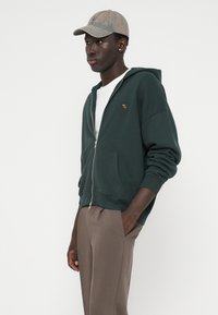 Haut à capuche zippé vert foncé avec de grandes poches avant, porté sur une chemise blanche. Assorti à une casquette gris clair avec un motif subtil et un emblème.