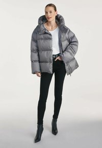 Szary pikowany puchowy płaszcz z wysokim kołnierzem i dużym kapturem, zestawiony z białą koszulą i czarnymi obcisłymi spodniami. Gładki, błyszczący materiał.