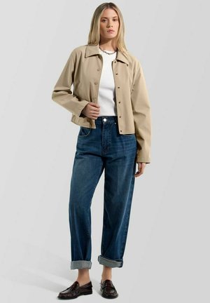 Frau trägt beige Knopfjacke, weißes Oberteil, hochgekrempelte blaue Jeans und dunkelbraune Loafers, steht vor einfarbig hellem Hintergrund.