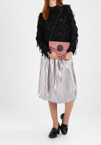 Bolso cruzado rosa con un acento de flor adornada, combinado con una blusa negra texturizada y una falda plateada metálica plisada, utilizado con zapatos negros.