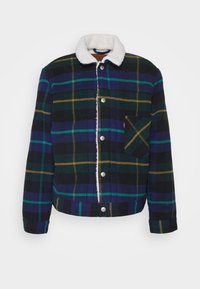 Chaqueta de franela con un patrón de cuadros en azul marino, verde y mostaza. Presenta un collar de pelaje sintético blanco y un bolsillo en el pecho. Botones de presión plateados.