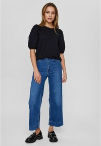 Svart kortärmad blus i texturerad tyg, kombinerad med vida blå denimjeans och svarta loafers. Modellen står på en neutral bakgrund.