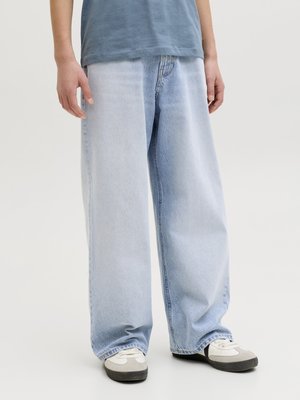 Inimene kannab laiade jalgadega, kahetoonilisi helesiniseid teksapükse, valgeid tossusid ja sinist T-särki, seistes ühevärvilise tausta ees.