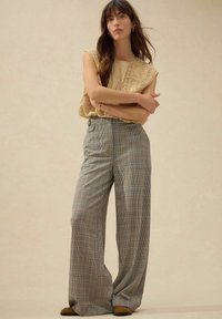 Haut en dentelle beige et pantalon large à carreaux bleu, gris et rouille, avec une taille haute et un motif à carreaux subtil. Des chaussures marron complètent le look.