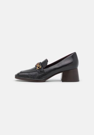 Mocassin en cuir noir à talon moyen, bout pointu et détail chaîne dorée sur le cou-de-pied, présenté sur un fond blanc.