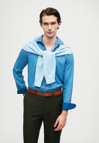 Pullover azzurro chiaro poggiato su una camicia di denim; cintura in pelle marrone; pantaloni verde scuro; texture morbide; design aderente.