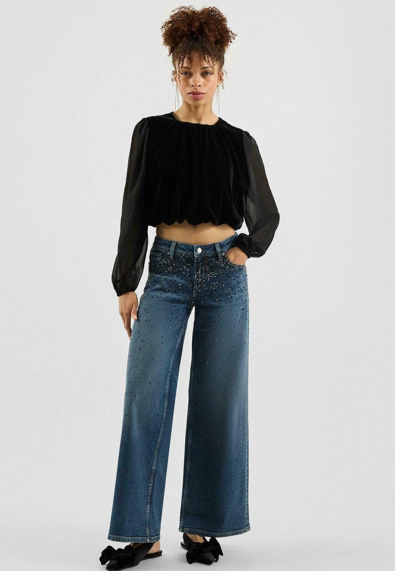 Zwart fluwelen cropped top met geplooid design, gecombineerd met wijde denim jeans met zilveren studs en zakken.