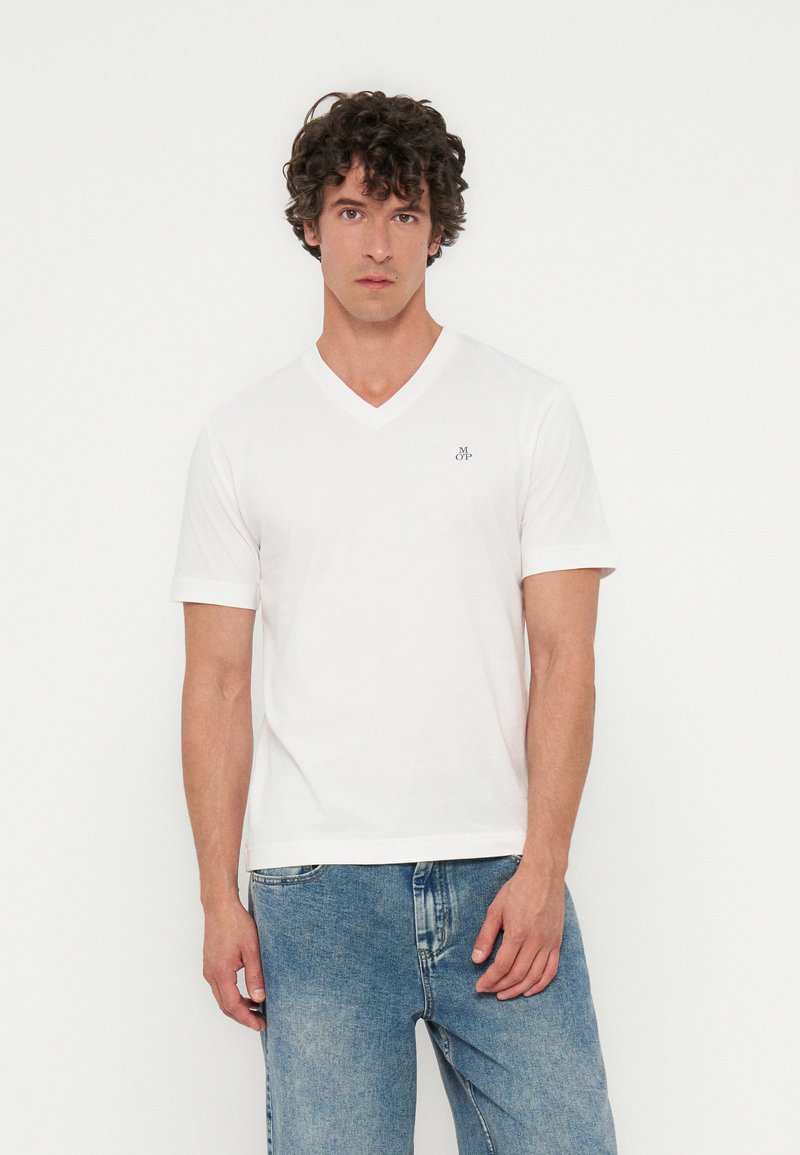 Άνδρας με κατσαρά μαλλιά, που φοράει λευκό T-shirt με V-λαιμόκοψη και μικρό λογότυπο στο στήθος και ανοιχτά μπλε τζιν, στέκεται μπροστά σε απλό φόντο.