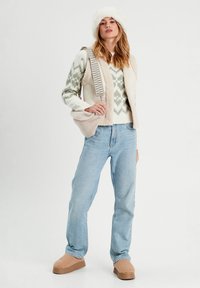 Ein cremefarbener und grüner gemusterter Pullover, eine beiges Fleece-Weste, hellblaue Jeans, tanfarbene, flauschige Slipper und ein weißer Pelzhut.
