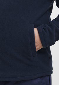 Jersey polar azul marino con una superficie texturizada, presenta un bolsillo lateral y puños ajustados. Incluye un pequeño logotipo en el dobladillo.