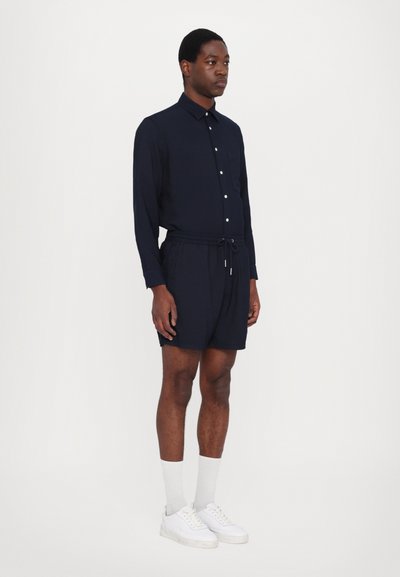 Marineblå skjorte med lange ermer og knapper, kombinert med marineblå trådføre korte bukser. Modellen har på seg hvite joggesko og lysegrå sokker.