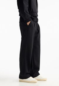 Personne portant un pantalon noir ample, une veste noire, les mains dans les poches, et des baskets blanches avec des semelles beiges, se tenant devant un fond blanc.