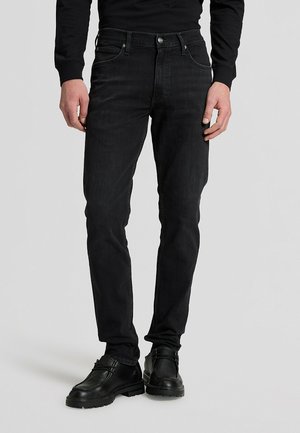 Persona che indossa jeans neri slim, camicia nera a maniche lunghe e scarpe nere con lacci, in piedi contro uno sfondo semplice.
