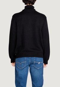 Maglione nero a maglia testurizzata con colletto alto, indossato su jeans in denim blu. Presenta polsini e orlo a coste, con cuciture visibili sui jeans.