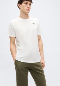 Άσπρο κοντομάνικο μπλουζάκι από λείο ύφασμα, με μικρό μαύρο λογότυπο Puma στο αριστερό στήθος. Συνδυάζεται με παντελόνι σε ελιά πράσινο χρώμα.