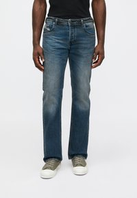 Homem com jeans azul desbotado de corte reto e ténis cinzentos, de pé contra um fundo branco liso.