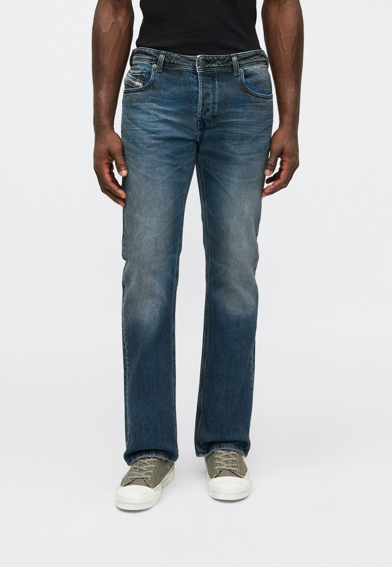 Homem com jeans azul desbotado de corte reto e ténis cinzentos, de pé contra um fundo branco liso.