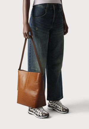 Rjava torba z vzorcem krokodila, z ravno dnom in nastavljivim trakom, v kombinaciji z ohlapnimi modrimi jeans hlačami ter sivo-rjavimi teniskami z belimi detajli.