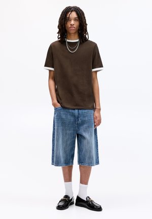 Bruine korte mouwen t-shirt met witte accenten, gecombineerd met lichtblauwe denim shorts. Zwarte instapschoenen, witte sokken en gelaagde zilveren kettingen.