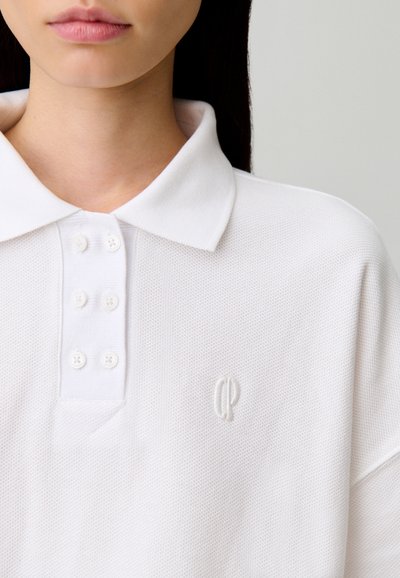Polo blanc en tissu texturé, avec un col classique, une patte de boutonnage à cinq boutons et un petit logo brodé sur la poitrine.