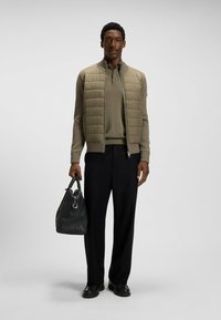 Puffer jacket in khaki, long-sleeved sweater in matching tone, black trousers. Man holds a black leather bag. Smooth textures, fitted design.