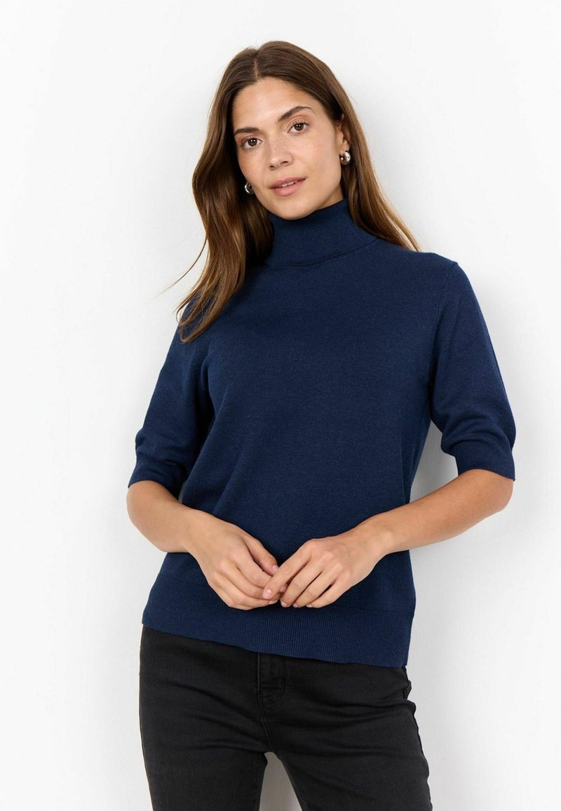 Pull turtleneck bleu marine à manches courtes en tissu doux. Présente un design ajusté et un ourlet côtelé. Porté avec un pantalon noir.
