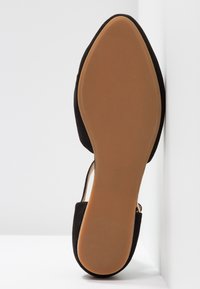 Chaussure plate noire à bout pointu avec semelle lisse marron clair, positionnée à la verticale contre un angle entre un mur et le sol blanc.