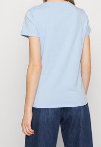 Persona vestida con una camiseta de manga corta azul claro y jeans de mezclilla azul oscuro, vista desde atrás sobre un fondo liso.