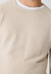 Beige pletený svetr s žebrovaným kulatým výstřihem a jemným texturovaným vzorem, který má dlouhé rukávy a přiléhavý střih.