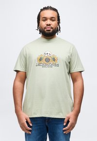 Άνδρας με πλεξούδες φοράει ανοιχτοπράσινο μπλουζάκι με σχέδιο κρανίου και ηλιοτρόπιων και μπλε τζιν, σε απλό φόντο.