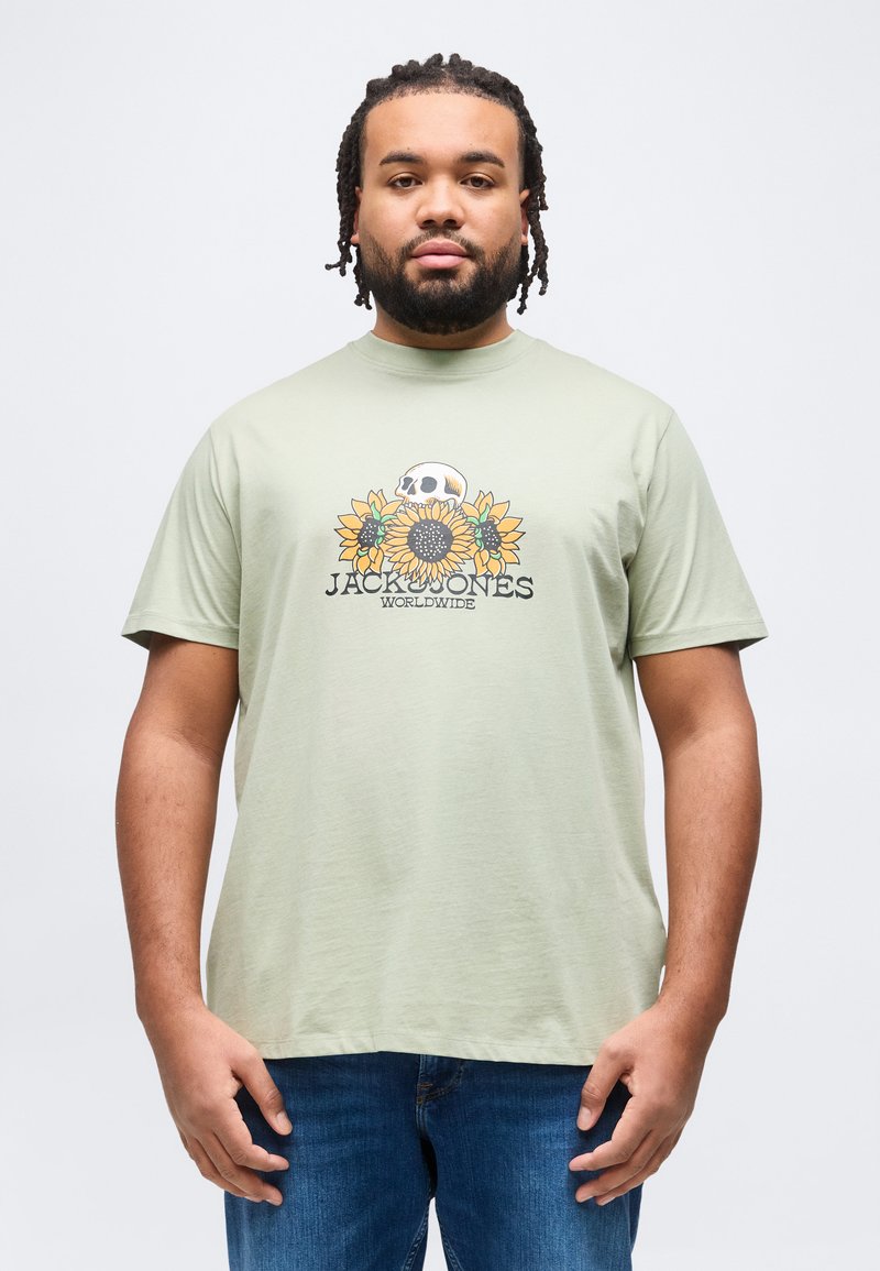 Άνδρας με πλεξούδες φοράει ανοιχτοπράσινο μπλουζάκι με σχέδιο κρανίου και ηλιοτρόπιων και μπλε τζιν, σε απλό φόντο.