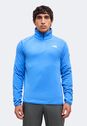 Άνδρας που φοράει φωτεινό μπλε fleece πουλόβερ North Face με μισό φερμουάρ, στέκεται μπροστά σε ένα απλό ανοιχτό φόντο.