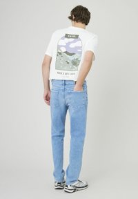 T-shirt blanc avec un graphique de montagne au dos, associé à un jean en denim bleu clair. Le design présente des verts, des violets et des accents de texte.