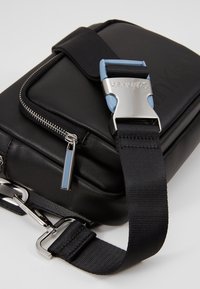 Czarna skórzana torebka na ramię z srebrnym zamkiem, metalową klamrą z niebieskimi akcentami z grawerem Calvin Klein oraz regulowanym czarnym paskiem.