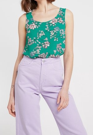 Woman wearing a green floral sleeveless top tucked into high-waisted light purple pants with front pockets and button closure.