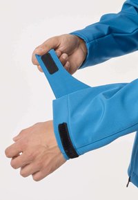 Mani che regolano la manica della giacca blu con chiusure a strappo nere, mostrando la cinghia di fissaggio tirata aperta.