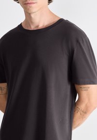 Tmavě šedé tričko s krátkým rukávem a kulatým výstřihem. Vyrobeno z hladkého materiálu, s malým logem umístěným v levém dolním rohu.