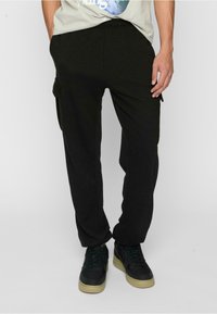 Black cargo pants with an elastic waistband, featuring side pockets. Worn with a light gray graphic t-shirt and dark sneakers.