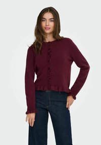 Pull en maille bordeaux avec un col rond, une fermeture à boutons à l'avant, un ourlet à volants et des détails aux poignets, associé à un pantalon en denim foncé.