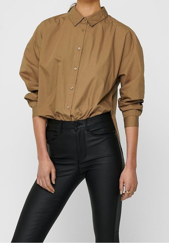 Brązowa koszula z guzikami, z kołnierzykiem, luźny fason i elastyczny dół, w połączeniu z gładkimi czarnymi skórzanymi spodniami. Wyraźny kontrast tekstur.