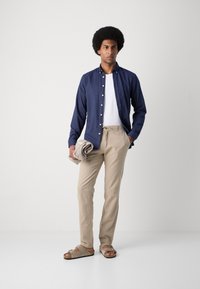 Navy button-up shirt, white T-shirt, beige drawstring pants, and light-colored sandals. Holding a folded gray item. Smooth textures, casual design.