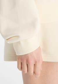 Puño de sudadera de canalé crema con textura suave, que presenta un dobladillo ajustado. Mano descansando sobre un muslo de piel clara en el fondo.