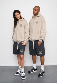 Sudaderas de cremallera beige con bolsillos frontales y logotipo, combinadas con pantalones cortos gris oscuro con logotipo. Ambos llevan zapatillas a juego y calcetines blancos.