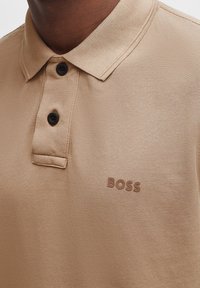 Beige poloshirt met een gestructureerde stof, drie donkere knopen en een subtiele bruine "BOSS" logo op de linker borst.